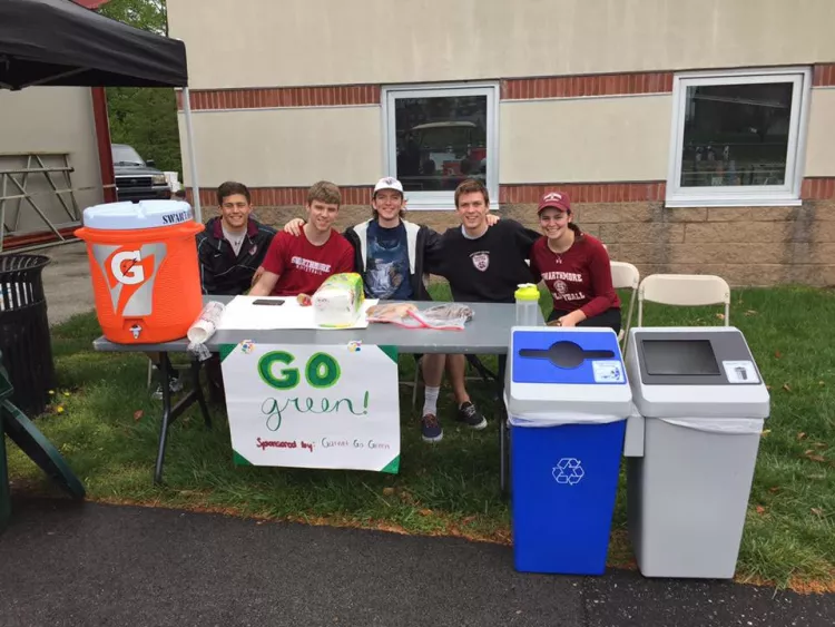 Swarthmore student athletes manning a table that is providing drinking water with compostable cups. A recycling bin next to a waste bin can be seen on the right, and a poster with the words "Go Green" is taped to the table.